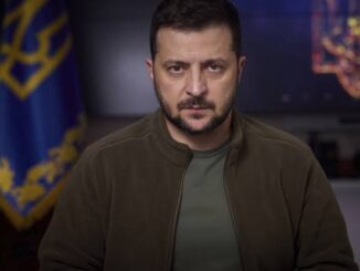 Man speaking in front of Ukrainian flag backdrop.