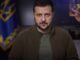Man speaking in front of Ukrainian flag backdrop.