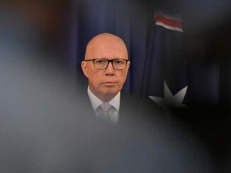 Bald man in suit and glasses before flag