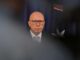 Bald man in suit and glasses before flag