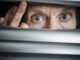 Person peering through window blinds, wide-eyed.