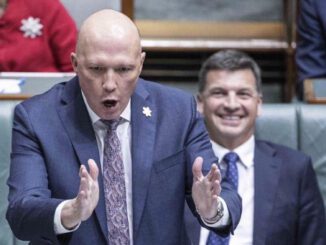 A man passionately speaking with hand gestures in a formal setting while another smiles in the background.