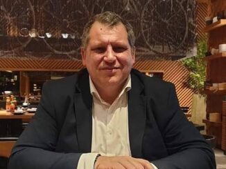 Man in a dark blazer and light shirt sitting at a table indoors.