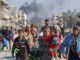 Crowd of people walking with smoke rising in the background.