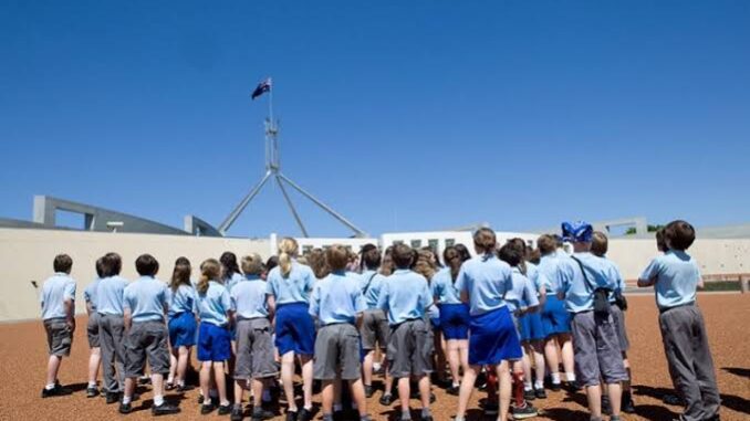 School children in uniforms gathered outside a government building under a clear blue sky.
