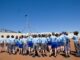 School children in uniforms gathered outside a government building under a clear blue sky.