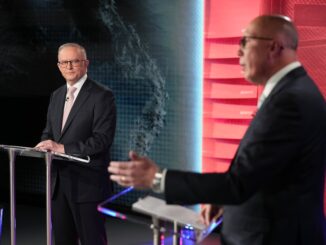 Two men engage in a formal debate or discussion on a stage with red and dark blue backgrounds.
