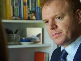 A serious man in a suit with bookshelves behind him.