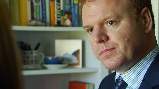 A serious man in a suit with bookshelves behind him.