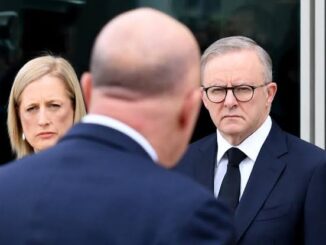 A serious man in glasses listens to a bald man speaking, with a woman observing.