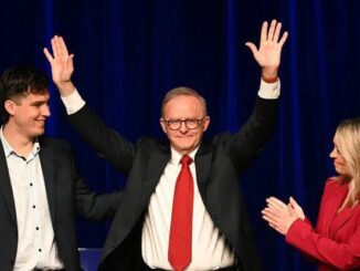 A man in a suit and red tie raising his hands in celebration on stage.