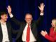 A man in a suit and red tie raising his hands in celebration on stage.