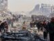 People walk through a large disaster site with rubble and debris.