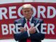 Former U.S. President Donald Trump posing with arms crossed in front of a sign.