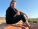 A person sitting on a rock with a smokestack in the background at sunset.