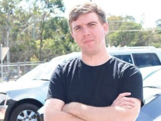 Man in black shirt with arms crossed standing outdoors.
