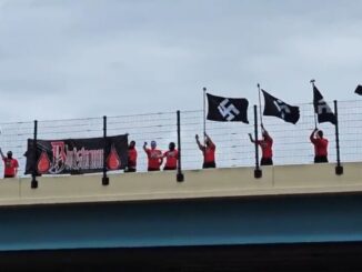 People on a bridge hold black flags and a banner under a cloudy sky.