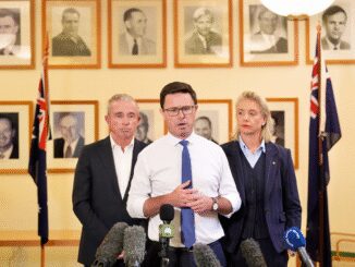 Three people standing at a press conference with framed portraits behind them.