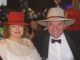 A smiling couple dressed elegantly with hats at a formal event.