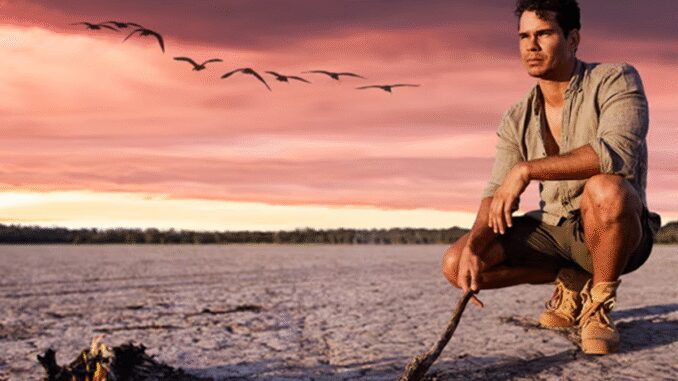 Man sitting by a lakeside during sunset with birds flying.