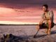 Man sitting by a lakeside during sunset with birds flying.