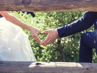 Bride and groom holding hands through a wooden fence outdoors.