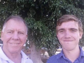 Two men smiling outdoors with greenery in the background.