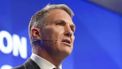 A man speaking with a microphone headset against a blue background.