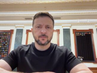 A man in a black shirt sitting indoors with ornate decor behind him.