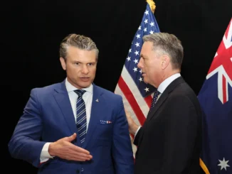 Two men in suits shaking hands in front of American flags.