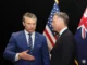 Two men in suits shaking hands in front of American flags.