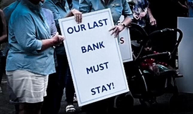 Protesters hold a sign saying 'OUR LAST BANK MUST STAY!'. Protesters hold a sign saying 'OUR LAST BANK MUST STAY!'.