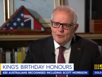 Man in glasses and suit being interviewed indoors with a framed flag in the background.
