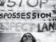 A person stands in front of a protest sign demanding respect for land and stopping dispossession.