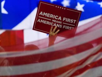 A hand holding a sign saying 'America First means Americans First' with a blurred American flag background.