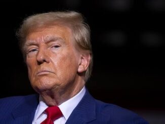 A serious man in a suit and red tie under dramatic lighting.