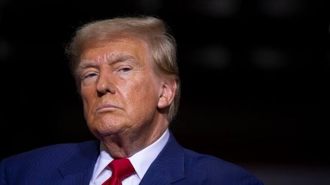A serious man in a suit and red tie under dramatic lighting.