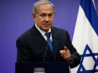 A man in a suit speaking at a podium with an Israeli flag in the background.