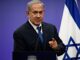 A man in a suit speaking at a podium with an Israeli flag in the background.