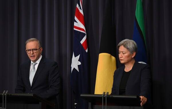 Two officials speak at a podium with Australian and Aboriginal flags behind them.