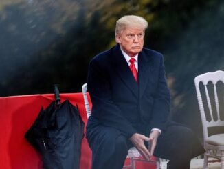 A man in a black coat and red tie sitting outdoors, looking thoughtful.