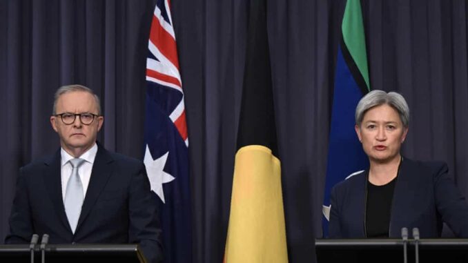 Two individuals seated at a table with Australian and Aboriginal flags in the background.