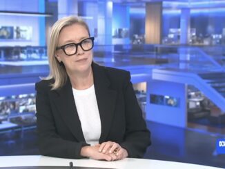 Woman in glasses and blazer sitting in a modern news studio.