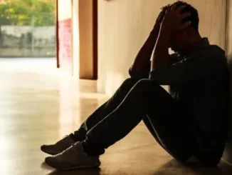 Person sitting against wall, holding head, distressed.