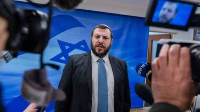 Man speaking to media with Israeli flag backdrop.