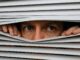 Person peeking through partially opened window blinds.