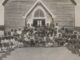 Large group of children outside wooden building.