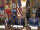Five officials seated beneath flags at table