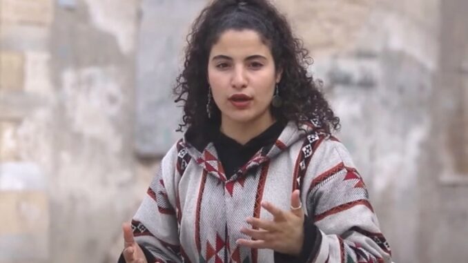 Woman speaking outdoors in patterned jacket.