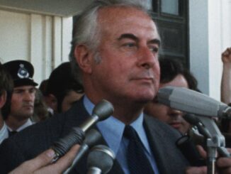 Man speaking at microphones, surrounded by crowd.